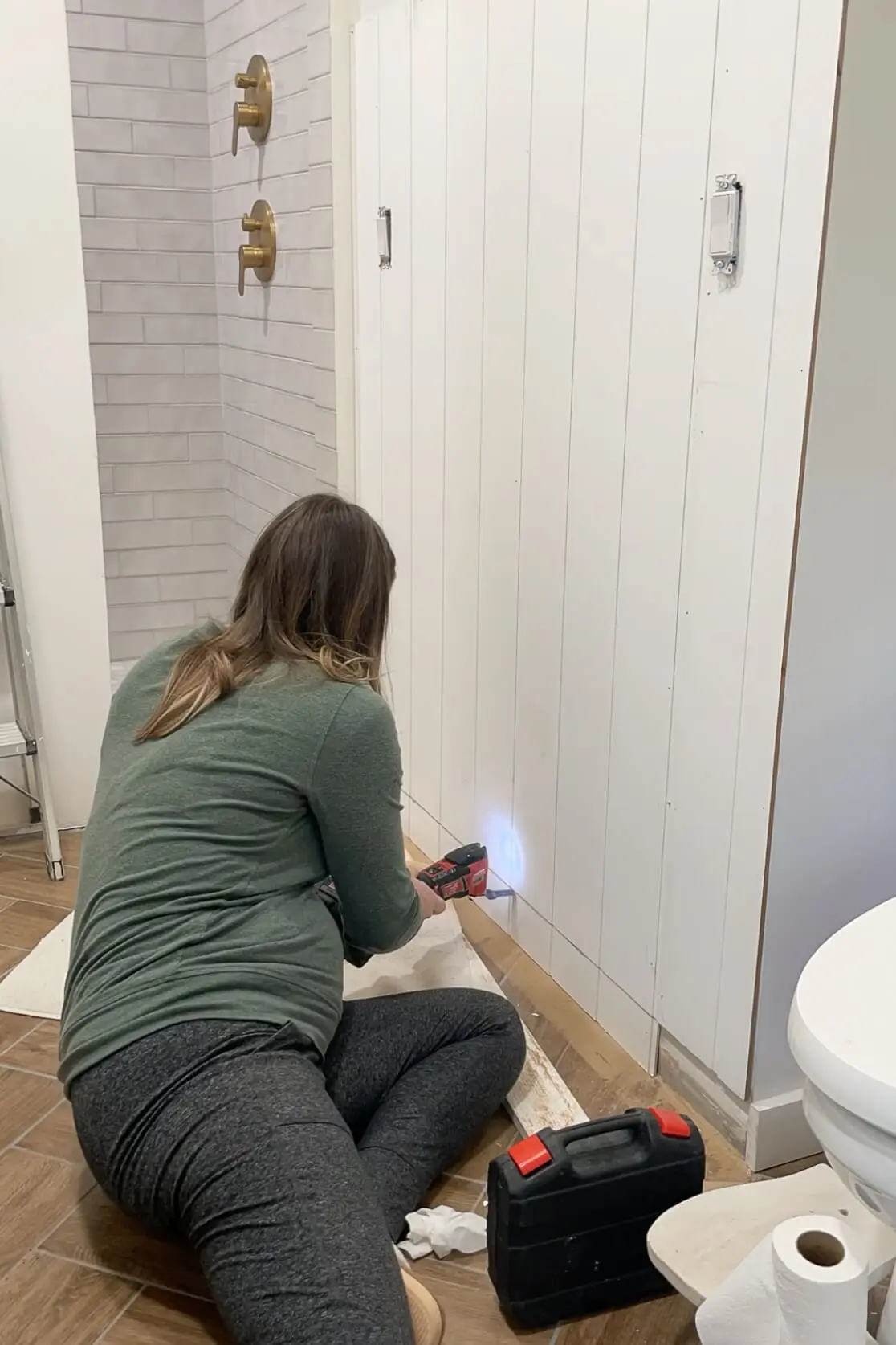 A woman kneeling on a brown herringbone tile floor, securing white vertical shiplap paneling to a bathroom wall with a power drill. The space features an arched shower entrance with gray subway tiles and brass shower fixtures. A tool bag and construction materials are nearby, showcasing the ongoing DIY renovation.